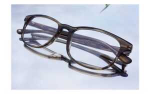 A pair of clear, rectangular eyeglasses with thin frames is placed on a reflective surface. The glasses have a subtle tint, and their reflection is visible beneath them.