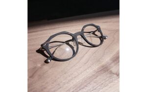 A pair of round, matte black glasses with clear lenses rests on a wooden surface. Bright lighting creates shadows and reflections, highlighting the frame and lens details.