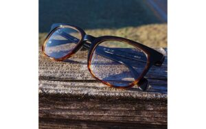A pair of glasses with brown and black frames rests on a wooden surface outdoors, with a blurred grassy area in the background. The sunlight casts a shadow of the glasses on the wood.
